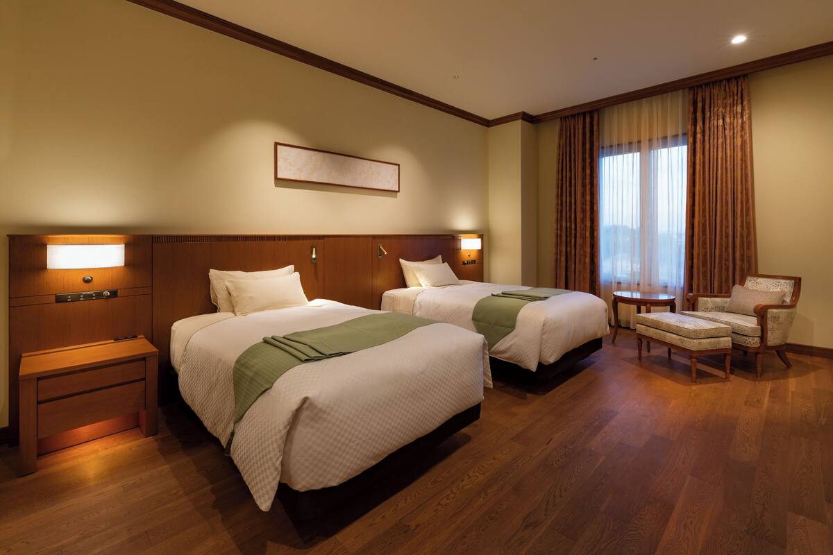 Een hotelkamer met twee bedden, een nachtkastje, een stoel en een groot raam met gordijnen.
