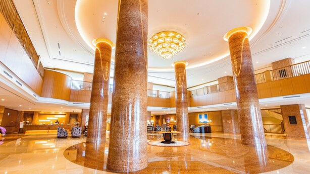 Un hall d’entrée spacieux avec de hauts plafonds, de grandes colonnes en marbre, un lustre central et un comptoir d’accueil.