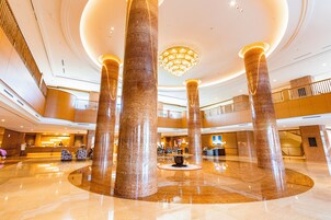 A spacious lobby with high ceilings, large marble columns, a central chandelier, and a reception desk.