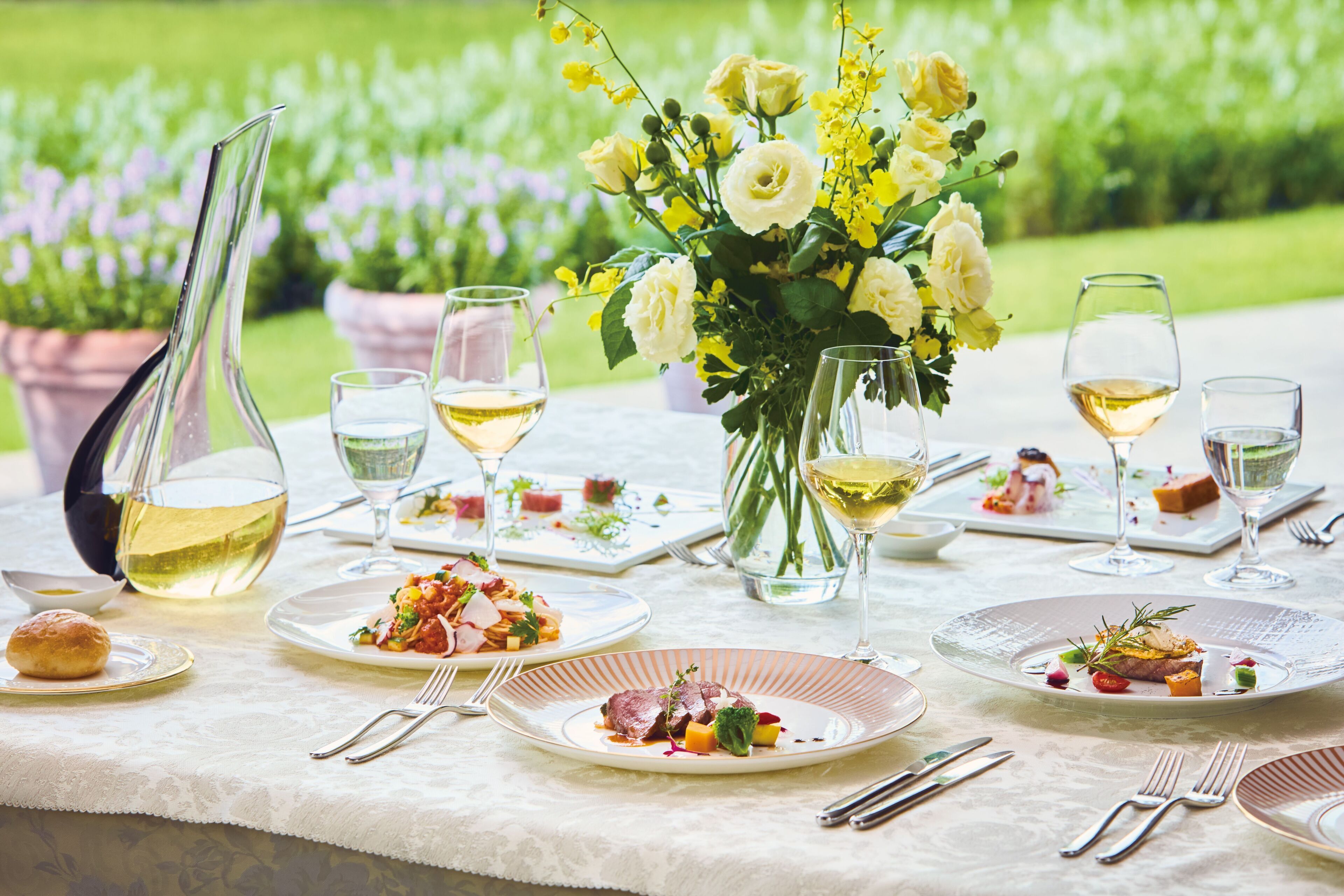 A table set for a meal with plates of food, glasses of white wine, and a vase of flowers.