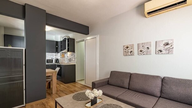 A modern living room with a grey sofa, a coffee table with decorative items, and a kitchen in the background. - Karavelov Top Center Apartment (Varna)