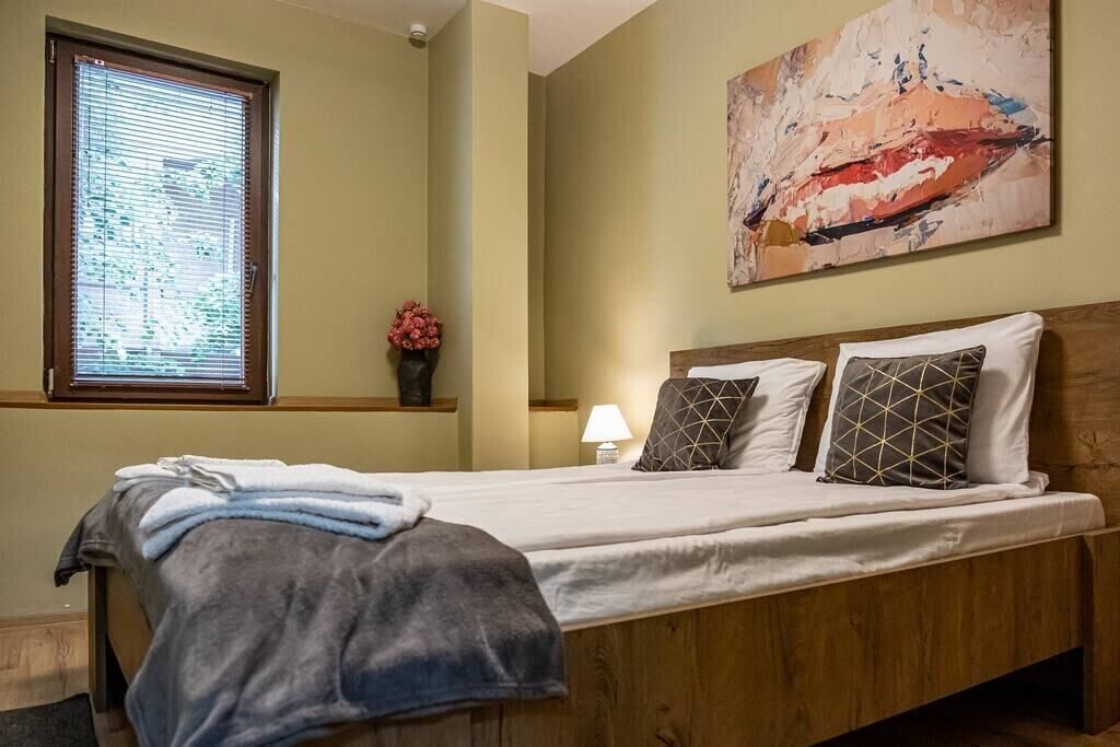 Une chambre avec un lit en bois, une fenêtre avec des stores, un tableau abstrait accroché au mur, et une petite table sur laquelle se trouvent une lampe et des fleurs.