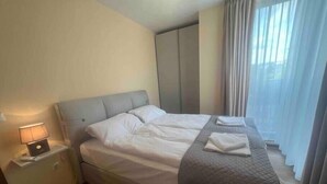 A hotel room with a bed, a nightstand with a lamp, a wardrobe, and a window with curtains. - Brand New Downtown Apartment (Varna)