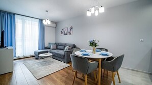 A modern living room with a grey sofa, a dining table with chairs, and a chandelier. - Brand New Downtown Apartment (Varna)