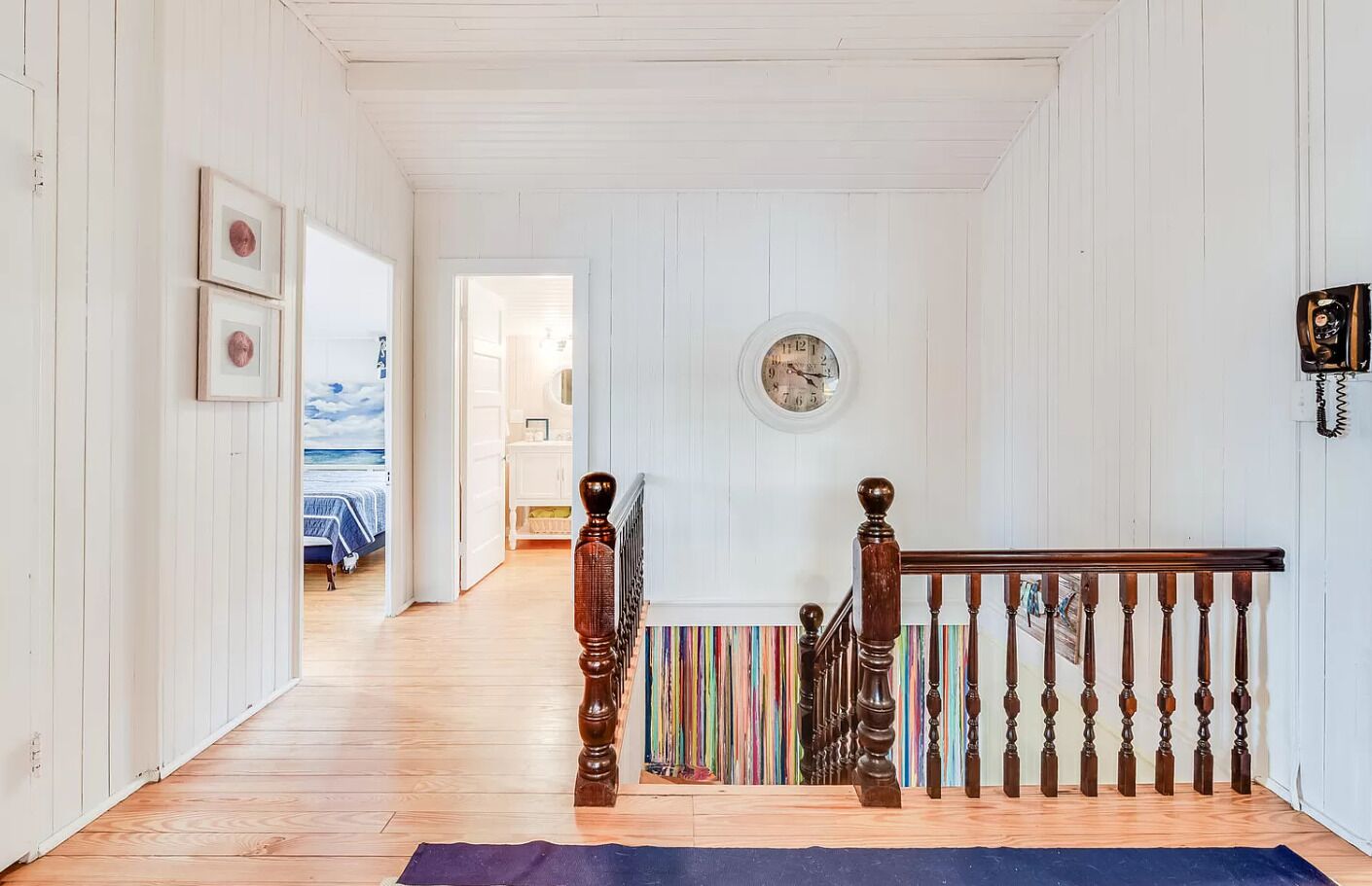 Un couloir au plancher de bois, un escalier recouvert d’un tapis à rayures et une horloge au mur.