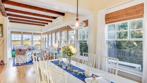 A dining area with a long table, white chairs, and a view of the outdoors. - Charming 6-bedroom oceanfront cottage in Falmouth (Falmouth)