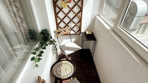 Un balcón estrecho con una silla de madera, una mesita redonda pequeña y una planta en maceta.