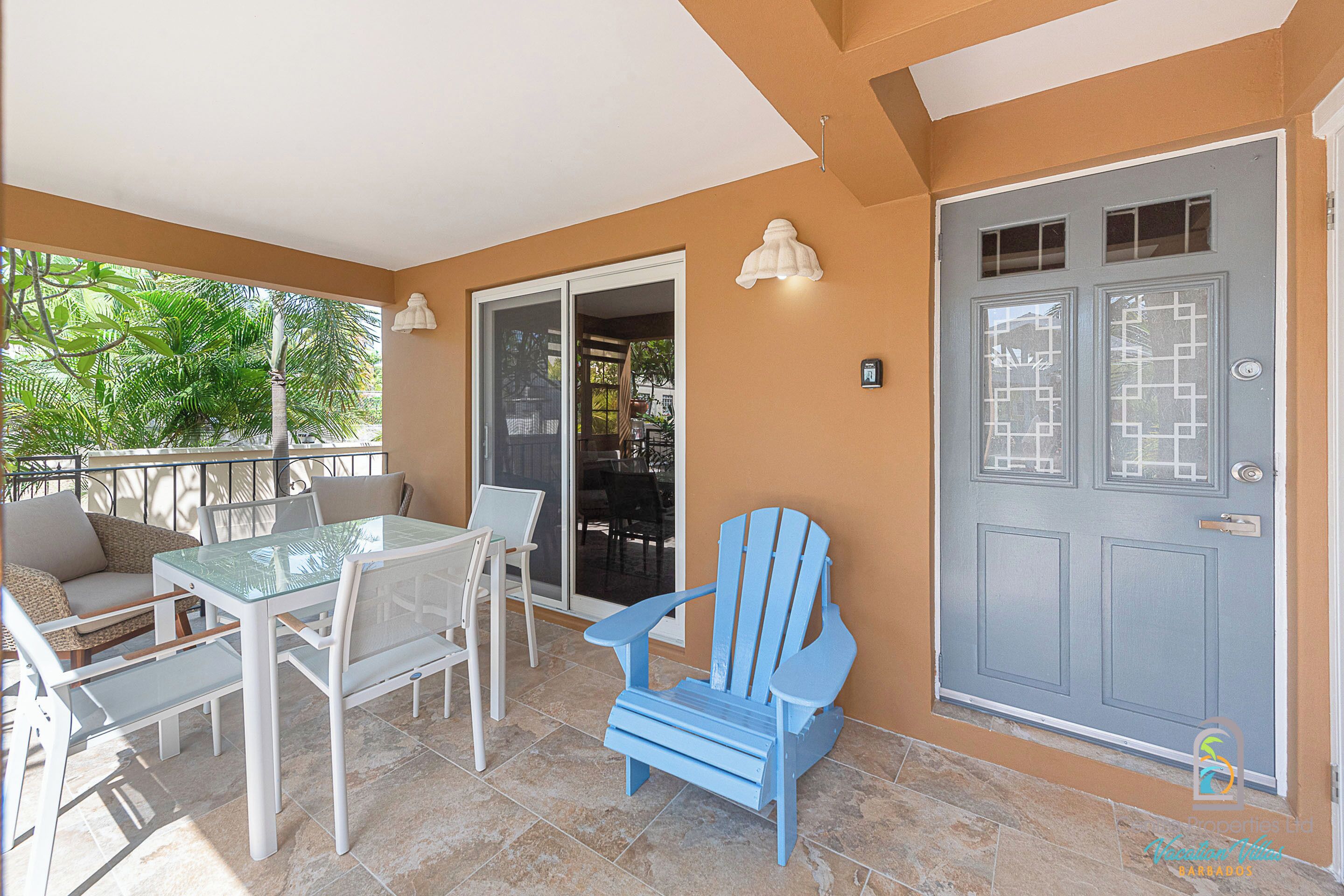 A balcony with a glass table, white chairs, a blue Adirondack chair, and a grey door with windows.