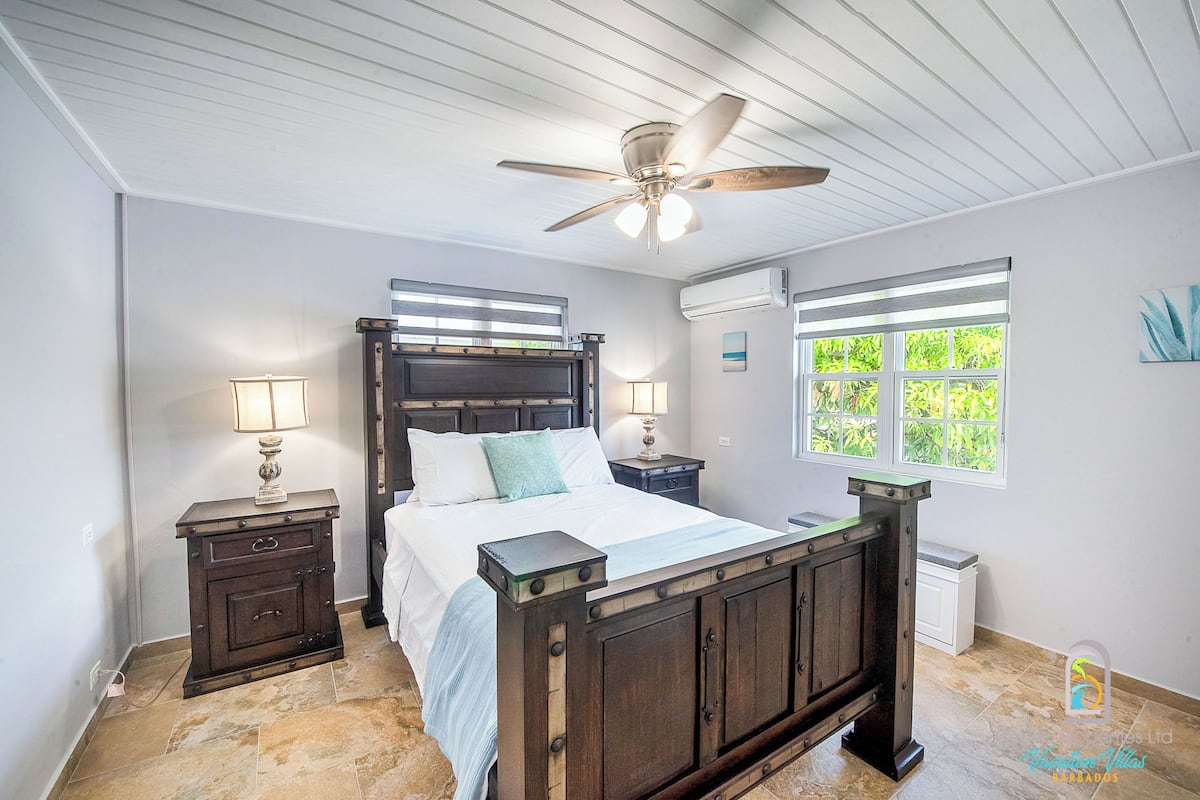 A bedroom with a wooden bed, two bedside tables with lamps, a ceiling fan, an air conditioning unit, and a window with a view of greenery.