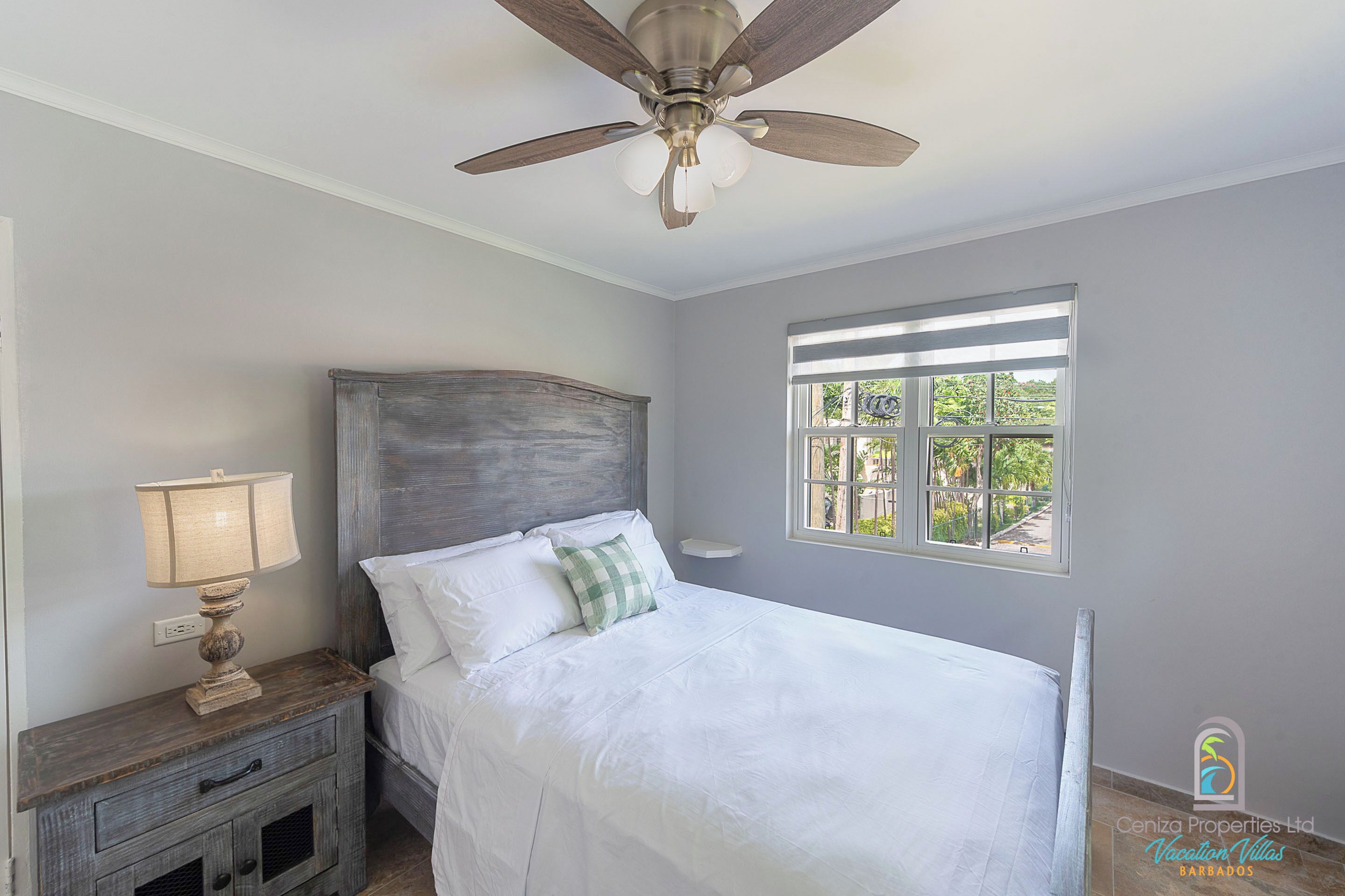 A bedroom with a bed, a nightstand, a lamp, a ceiling fan, and a window with a view of greenery.