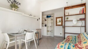 Ein Zimmer mit Esstisch, Stühlen, einem Bett mit bunter Decke, einem Bücherregal, einem Spiegel und einem Heizkörper.