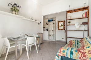 Une pièce comprenant une table à manger, des chaises, un lit avec une couverture colorée, une bibliothèque, un miroir et un radiateur.