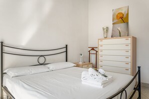 A neatly made bed with white linens, a wooden nightstand with a lamp, a dresser with drawers, and a framed picture on the wall.