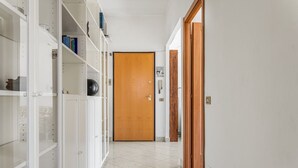 Un couloir avec une porte en bois au fond, des étagères blanches sur le côté gauche et un sol carrelé.