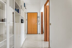 Un couloir avec une porte en bois au fond, des étagères blanches sur le côté gauche et un sol carrelé.