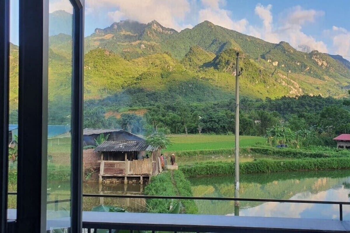 Une vue depuis une fenêtre sur un paysage rural, avec des montagnes, un étang et des maisons traditionnelles.