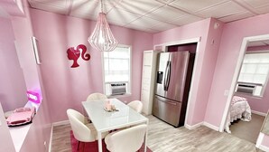 A pink-themed room with a dining table, chairs, a chandelier, a refrigerator, and a window with blinds. - The Dollhouse  (Old Orchard Beach)