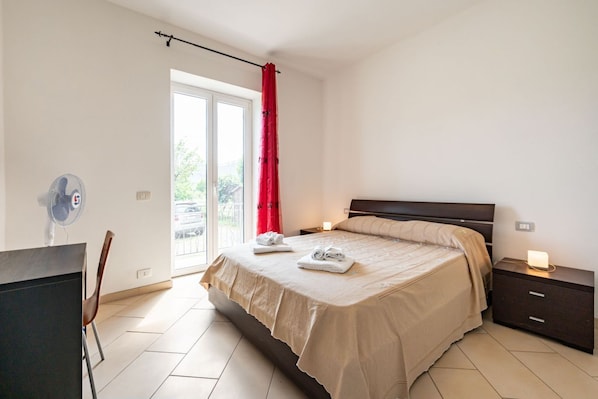 A hotel room with a bed, a desk, a chair, a nightstand, and a window with a red curtain.