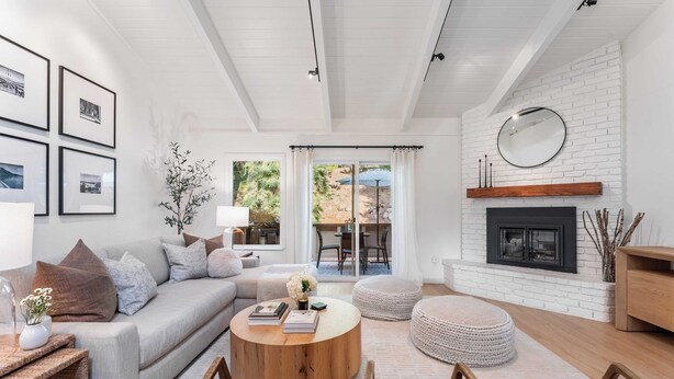 Una sala de estar moderna con sofá, mesa de centro y reposapiés, chimenea y zona de comedor visible a través de un vidrio.