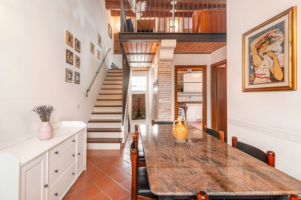 A dining area with a marble table, wooden chairs, a staircase, and a kitchen in the background.