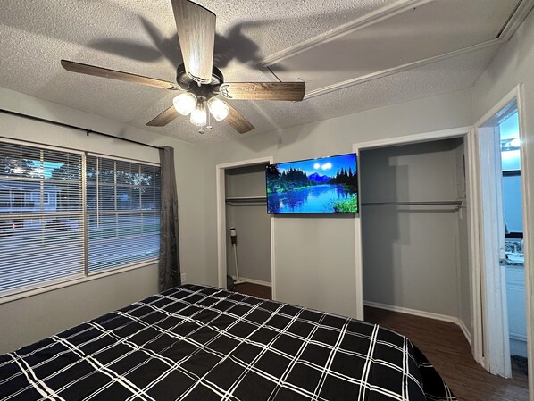 Un dormitorio con cama, ventilador de techo, televisor montado en la pared y ventana con persianas.