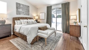 Une chambre avec un grand lit, une table de chevet, une commode, un ventilateur de plafond, une fenêtre avec des rideaux et une vue sur l’extérieur.