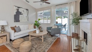 Un salon moderne avec un canapé blanc, un fauteuil gris et une table basse ronde. On y trouve un ventilateur de plafond, une cheminée et un grand tableau abstrait accroché au mur.
