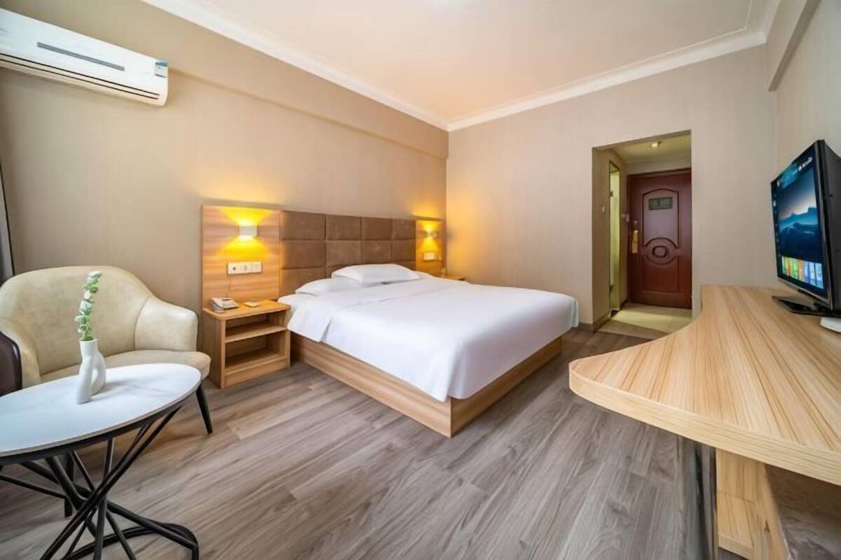 Ein Hotelzimmer mit Bett, Nachttischen, einem Sessel, einem kleinen Tisch, einem Fernseher und einer Klimaanlage.