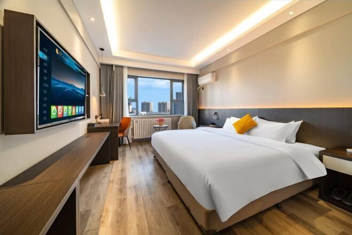 Ein modernes Hotelzimmer mit einem großen Bett, einem Schreibtisch, einem an der Wand befestigten Fernseher und Blick auf die Stadt durch das Fenster.
