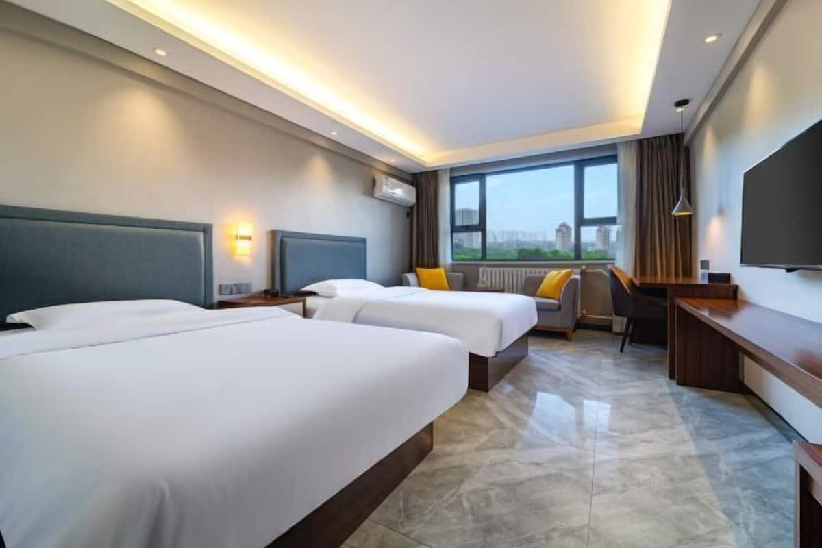 Ein Hotelzimmer mit zwei Betten, einem Schreibtisch, einem Stuhl, einem Fernseher und einem Fenster mit Blick auf die Stadt.