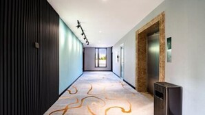 A modern hotel hallway with a patterned carpet, a trash bin, and a wall-mounted light fixture.