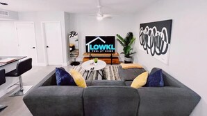 A modern living room with a grey sofa, a coffee table, and a TV screen displaying 'LOWK'L FEEL AT HOME'. - Bright 3-Bedroom 3 Bath Apt with Pool, Parking, & Close to Beach & Las Olas (Fort Lauderdale)