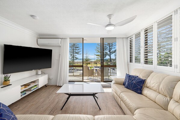 A living room with a flat-screen TV, a white coffee table, a beige sofa with blue pillows, and a balcony with a view of the beach and trees. - Beachfront Beauty - Cooly (Gold Coast)