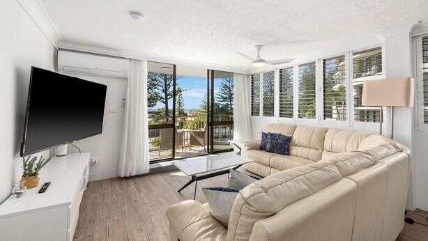 A modern living room with a flat-screen TV, a beige sofa, a glass coffee table, and a ceiling fan. - Beachfront Beauty - Cooly (Gold Coast)