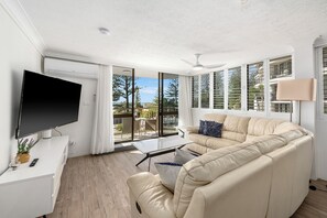 A modern living room with a flat-screen TV, a beige sofa, a glass coffee table, and a ceiling fan. - Beachfront Beauty - Cooly (Gold Coast)