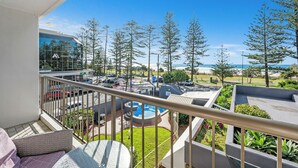 A balcony with a view of a beach, a pool area, and a building with a sign. - Beachfront Beauty - Cooly (Gold Coast)