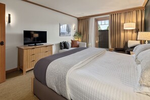 Ett hotellrum med en stor säng, en tv placerad på ett träskåp, ett skrivbord med en lampa och utsikt över omgivningarna genom en skjutdörr.