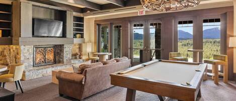 A spacious room with a pool table, comfortable seating, a fireplace, and large windows offering a mountain view.