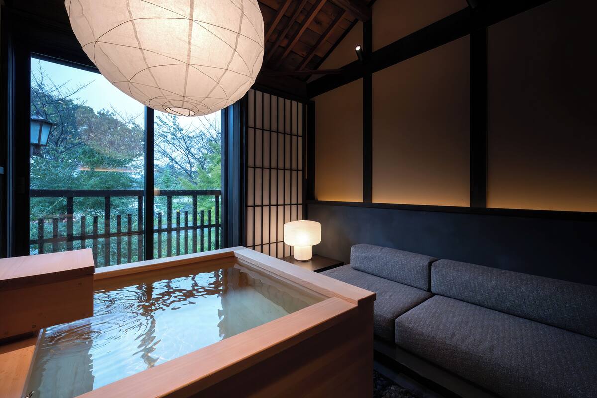 Una habitación japonesa tradicional con suelo de tatami, un baño de aguas termales de madera, un sofá y un ventanal con vistas a un jardín.