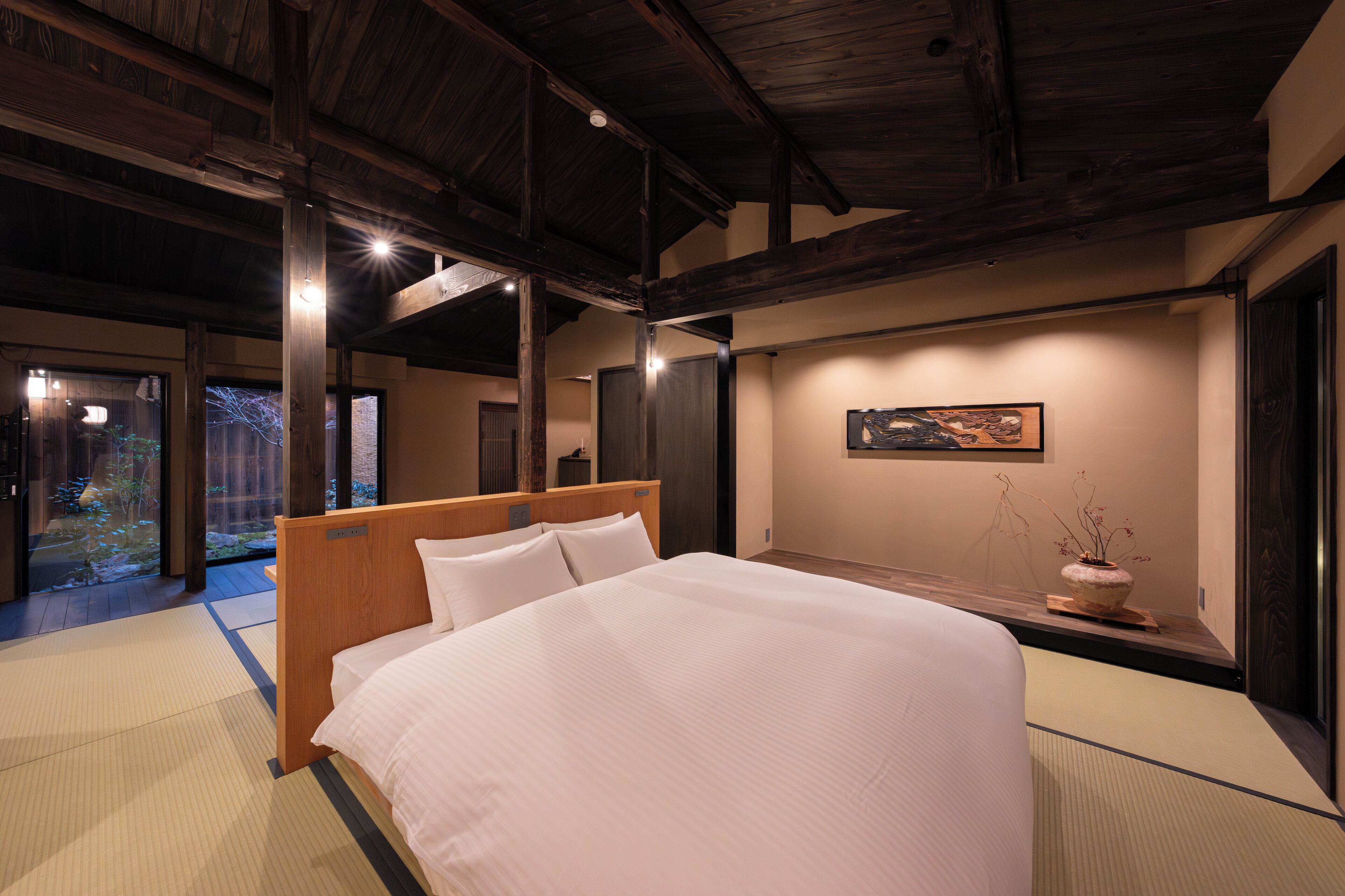 A hotel room with a large bed, wooden headboard, tatami mat flooring, a view of a garden, and a painting on the wall.