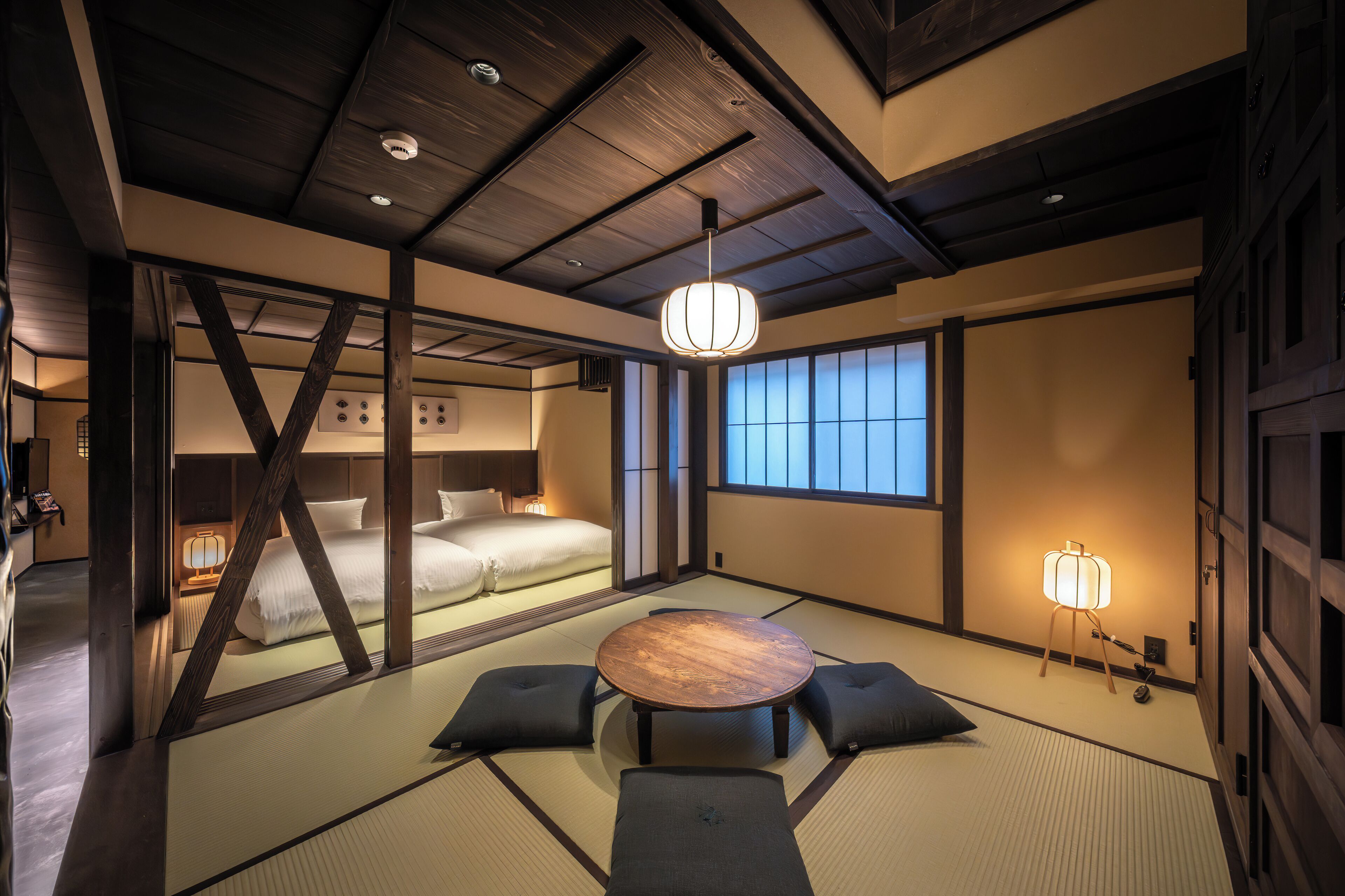 Una habitación japonesa tradicional con suelo de tatami, una mesa baja con cojines, una cama con edredón blanco y una puerta corrediza que da a otra habitación.