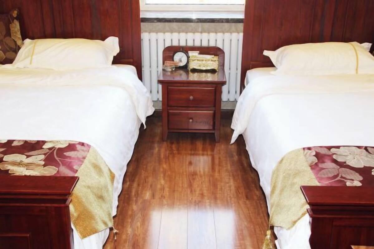 Een hotelkamer met twee enkele bedden, een houten nachtkastje met een klok en boeken, en een radiator op de achtergrond.