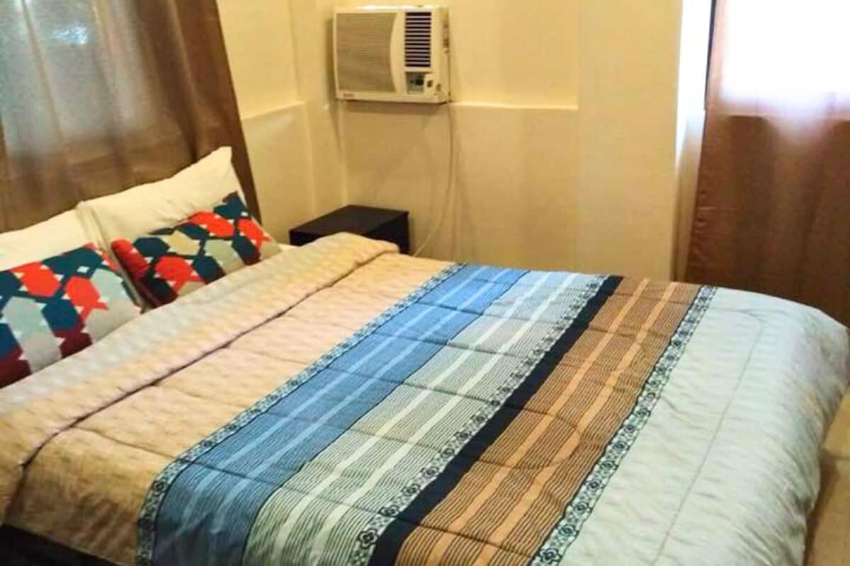 Une chambre d’hôtel comprenant un lit, une table de chevet, un appareil de climatisation et une fenêtre avec des rideaux.