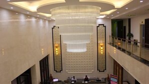 Um saguão de hotel com uma mesa de recepção, um lustre grande e um corrimão de vidro no nível superior.