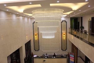 Um saguão de hotel com uma mesa de recepção, um lustre grande e um corrimão de vidro no nível superior.