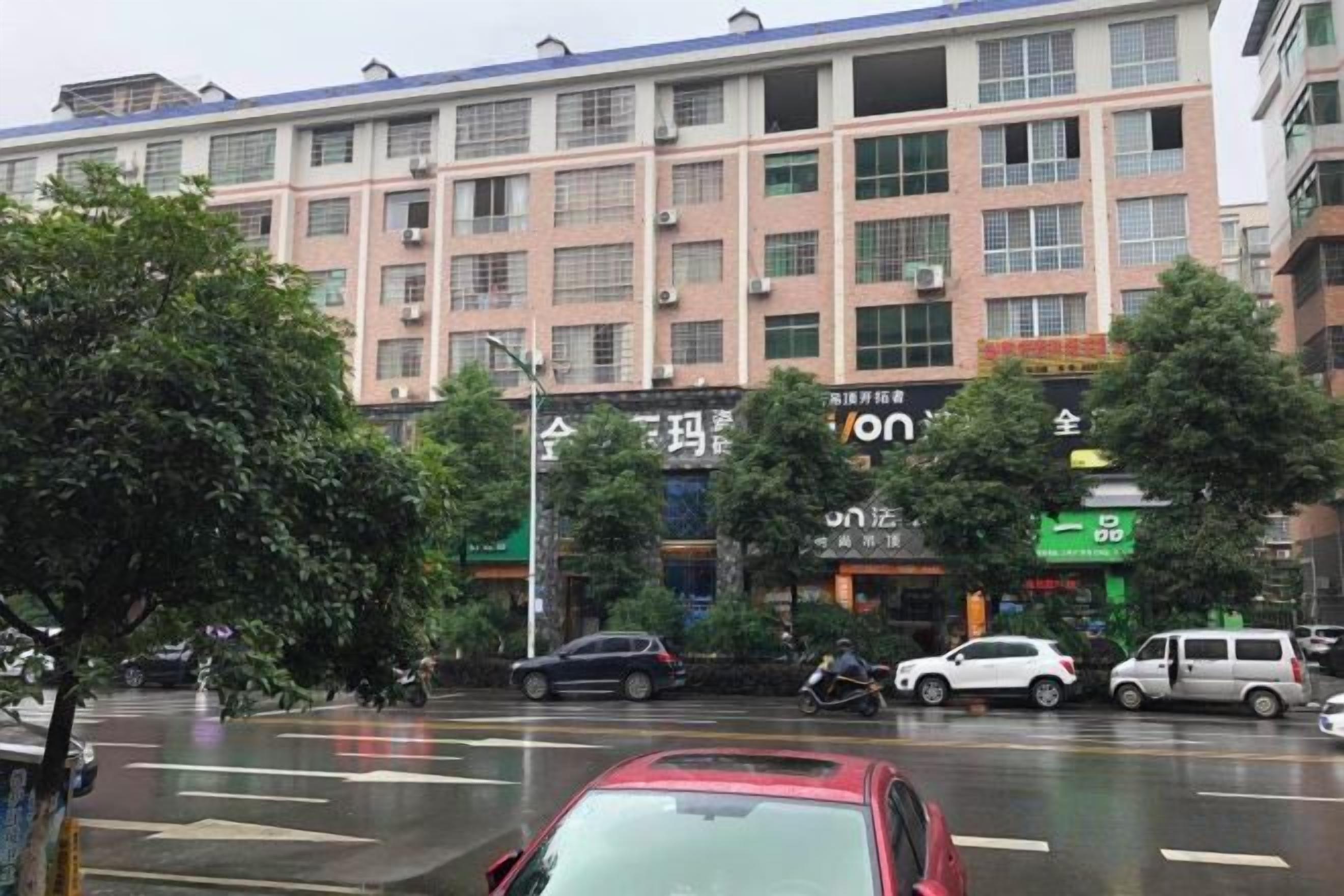 A multi-story building with commercial establishments on the ground floor, a wet street, and vehicles parked along the curb.