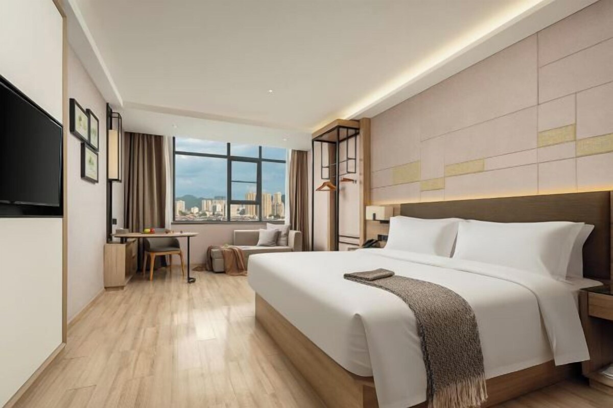 A hotel room with a large bed, a desk, a chair, a TV, and a window with a city view.