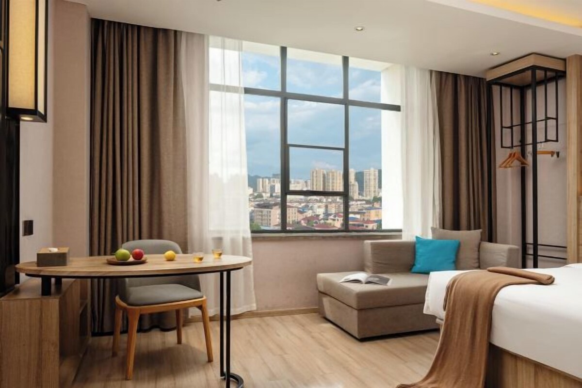 A hotel room with a large window, a bed, a sofa, a table with fruits, and a chair.