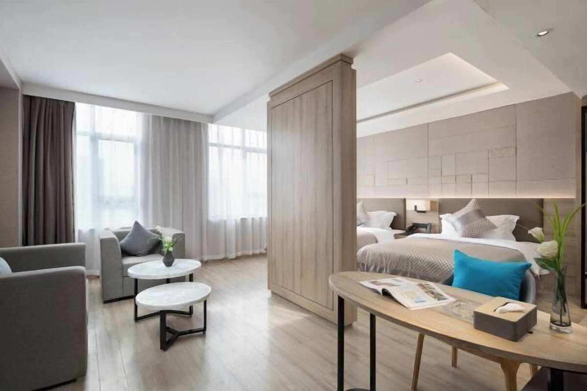 A modern hotel room with a large bed, a sitting area with a sofa and two round tables, and a large window with curtains.
