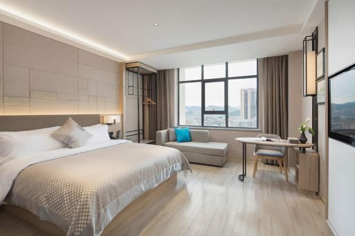 A modern hotel room with a large bed, a sofa, a desk, and a view of the city.
